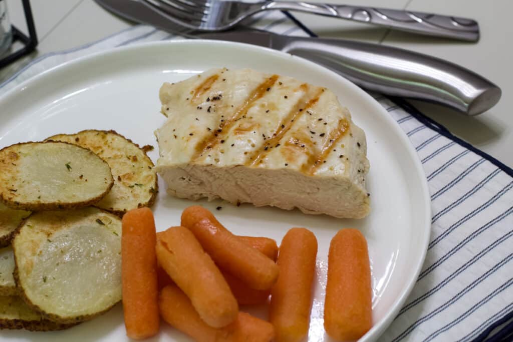 One half of a cooked chicken breast on a plate with air fried potatoes and cooked baby carrots.
