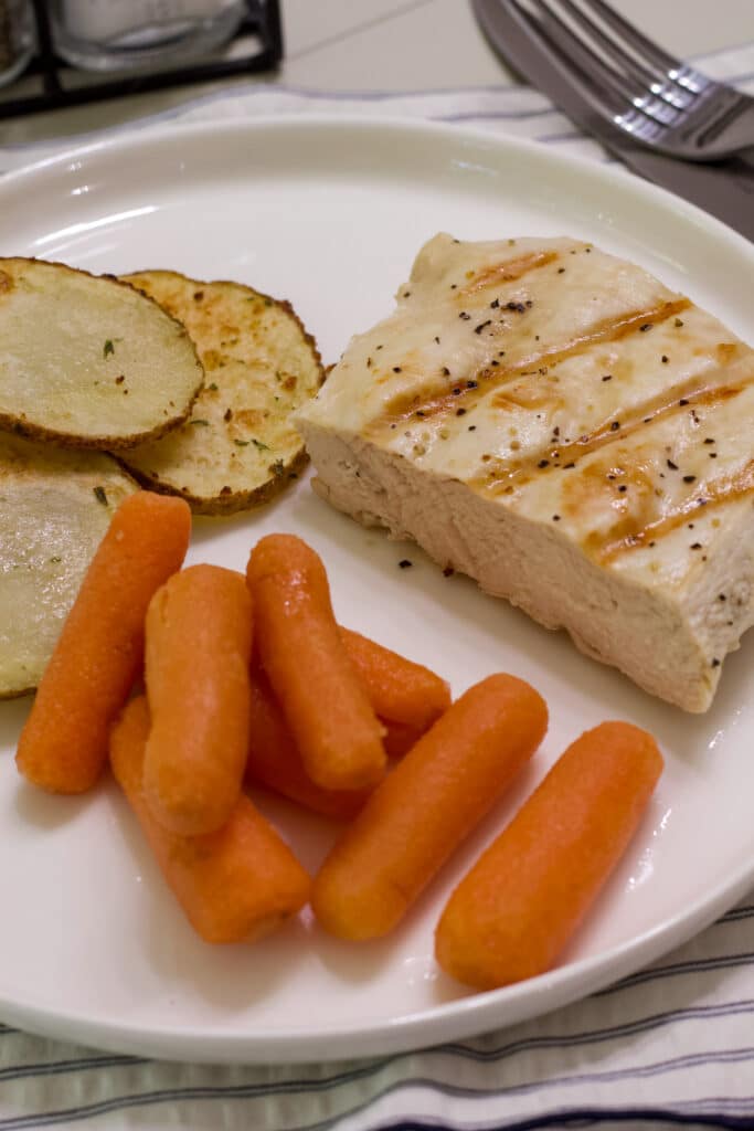 One half of a cooked piece of boneless chicken on a plate with air fryer potatoes and cooked baby carrots.
