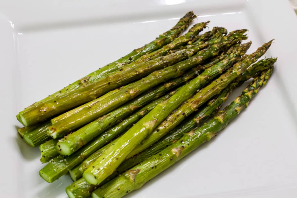 Many asparagus spears after they were roasted in a 350° oven, on a white serving plate.