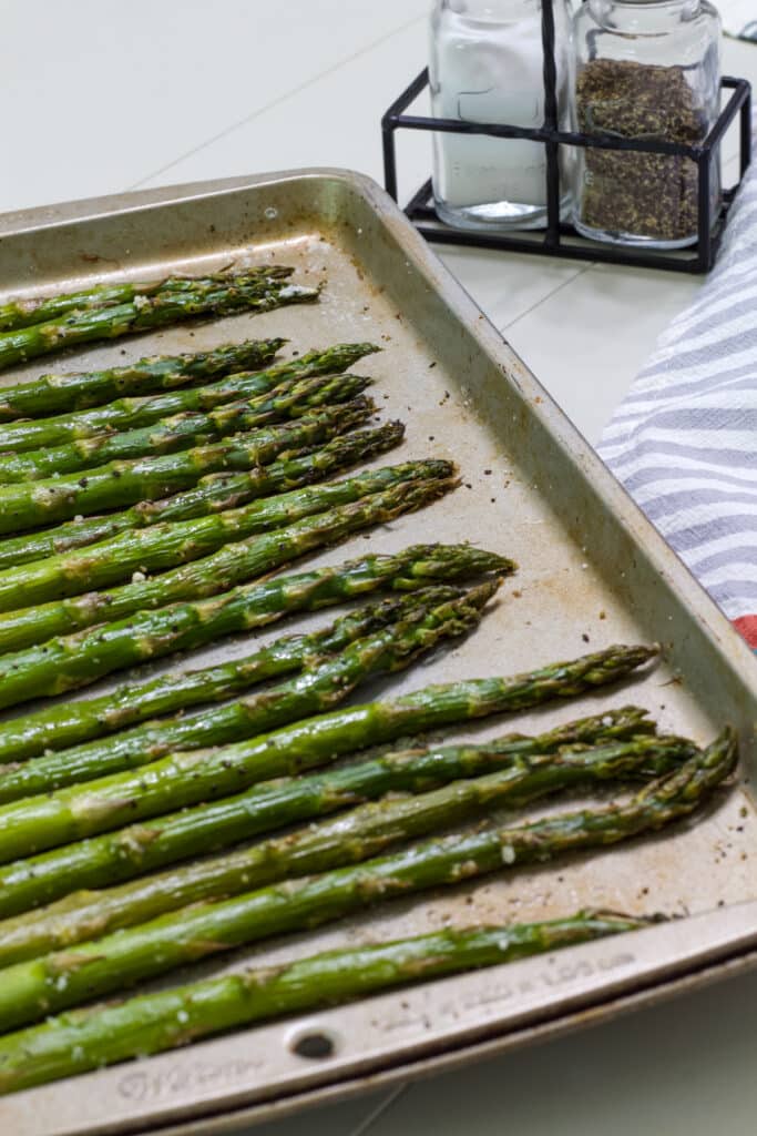 A rimmed baking sheet with oven-roasted asparagus on it in the foreground and salt and pepper shakers in the background.