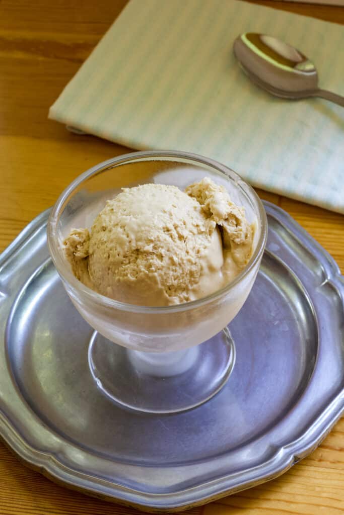 One scoop of ice cream in a glass bowl that is sitting on a silver plate.
