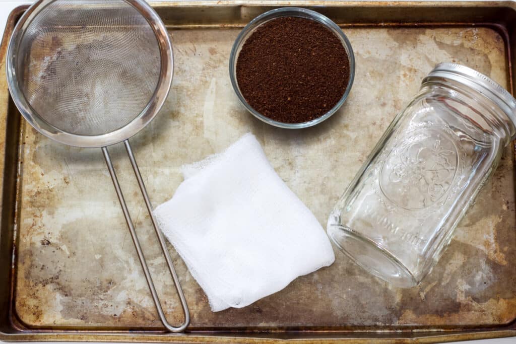 A rustic sheet pan with a mesh strainer, cheese cloth, small bowl of ground coffee and a large mason jar on it to make the cold brew coffee with.