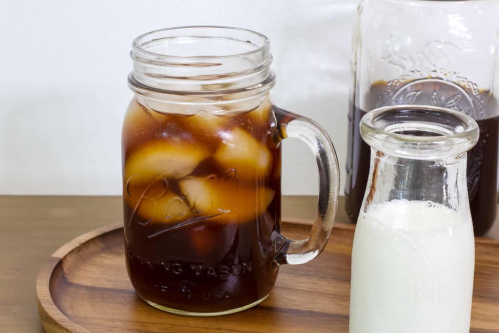 A mug of iced cold brew coffee and a small bottle of milk, there is a mason jar of coffee in the background.