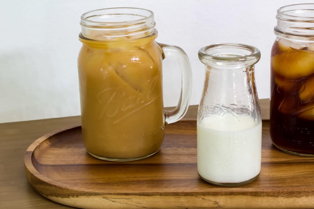 A mug of coffee with milk in it, a small bottle of milk and part of another mug of iced cold brew is to the right of the milk.